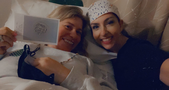 Two women lie close together smiling; one holds a photo of a cluster of cells, possibly an embryo, and the other wears a patterned cap.